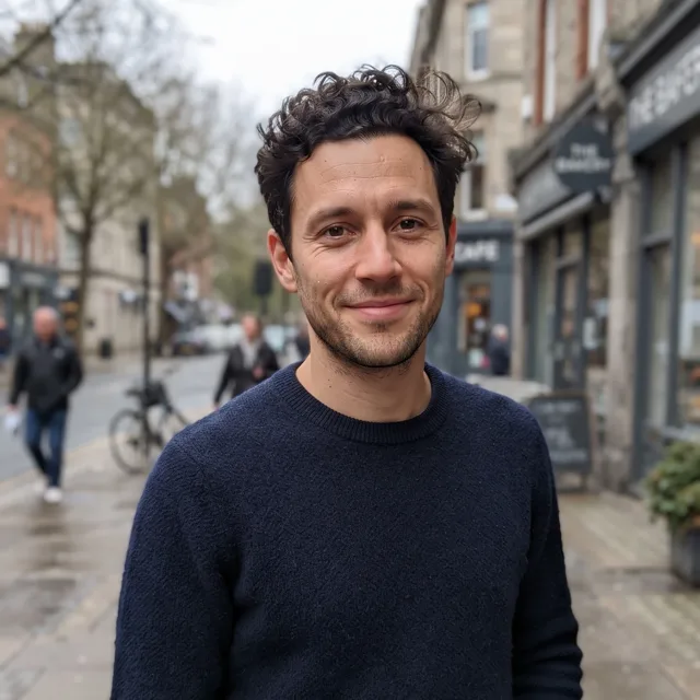 A realistic everyday photo of a man in his late twenties with short dark curly hair and light stubble, wearing a navy sweater on an overcast city street and smiling slightly, the starting photo for a cyberpunk portrait.