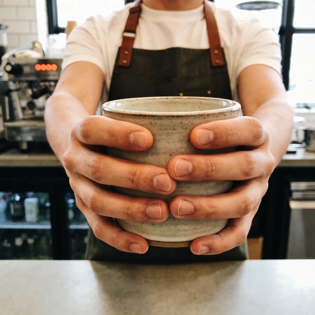 A barista in a dark apron holds a ceramic cup out toward the camera with both hands, fingers wrapped around the sides, against a blurred espresso machine in the background.