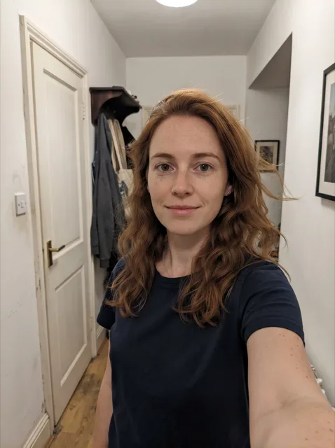 An ordinary phone selfie of a young woman with wavy auburn hair and freckles in a navy t-shirt, standing in a narrow apartment hallway with a white door and a coat rack under flat ceiling light.