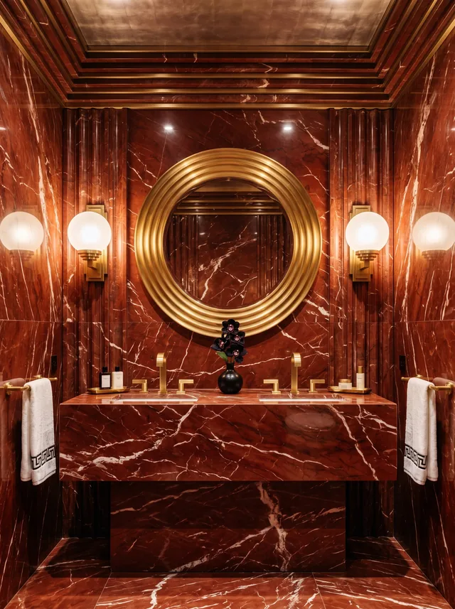 Art deco bathroom clad in red marble with a slab vanity, round brass-ringed mirror and globe sconces