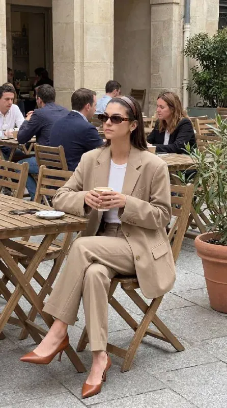 Photo de Seedream d'une femme en costume beige ample et lunettes de soleil écaille assise avec un café à la terrasse d'un café en plein air, avec d'autres clients flous derrière elle