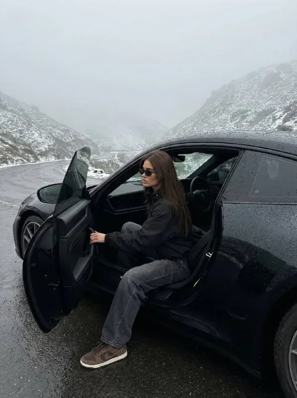 زنی با کاپشن مشکی و شلوار جین گشاد پشت فرمان پورشه 911 مشکی روی جاده کوهستانی خیس و مه‌آلود نشسته، از نمای نیم‌رخ