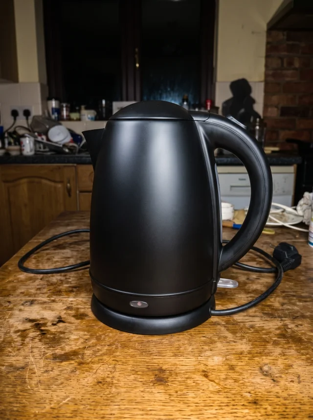 Matte black electric kettle on a wooden table, cleaned up from a dim, dusty, yellow-cast photo into a bright, sharp, colour-corrected product shot with the same angle and background