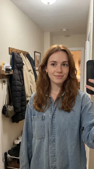 An ordinary vertical mirror selfie of a young woman with wavy auburn hair and freckles in an oversized denim shirt, standing in a narrow apartment hallway with coats on hooks behind her.