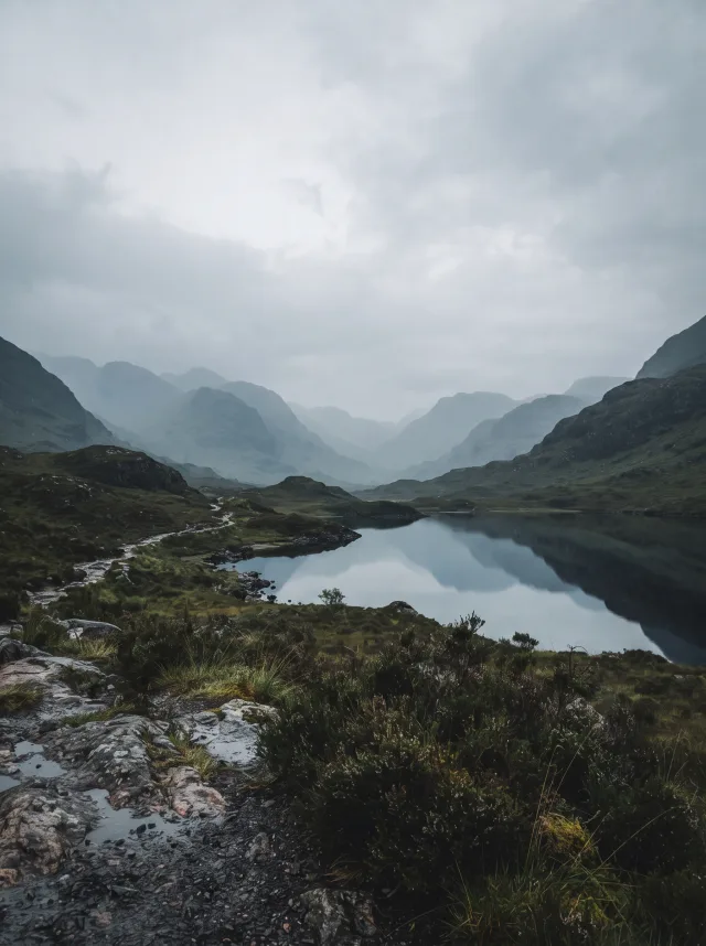 Misty Scottish highland glen at dawn with layered ridges, a dark loch and heather in slate and moss tones